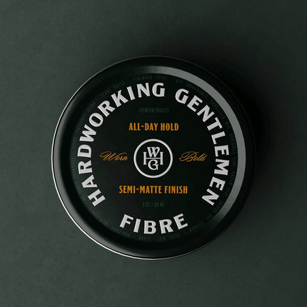 A round black tin of WG Haircare Hardworking Gentlemen Fibre All-Day Hold, shown from a top-down view on a dark charcoal background.